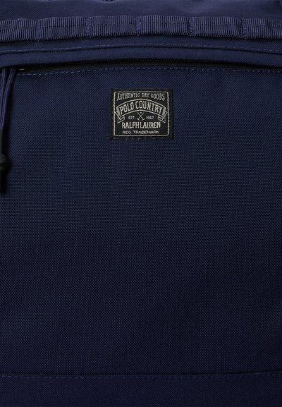 Υφασματινή υφή ναυτικού μπλε με ραμμένη ορθογώνια ετικέτα Polo Country Ralph Lauren, πάνω από μια τσέπη με φερμουάρ.