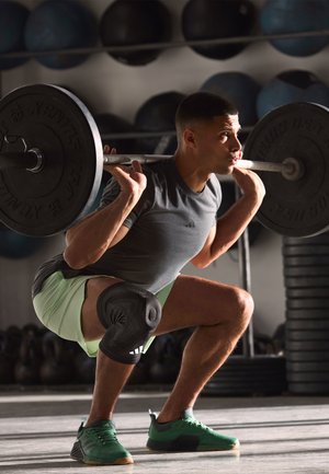 Black weightlifting bar with plates supported on shoulders; wearing a gray shirt, green shorts, and green sneakers; black knee support featured.