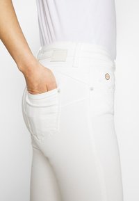 Jeans de denim blanco con un corte ajustado, que presentan un bolsillo trasero, costuras claras y una etiqueta de marca en la cinturilla. Textura suave.