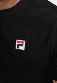 Black cotton t-shirt featuring a small embroidered rectangular logo with red, navy, and white colors on the chest. Round neckline.