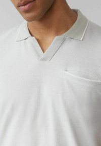 Hellgraues Polohemd mit kurzen Ärmeln und V-Ausschnitt, mit strukturiertem Material und einer kleinen Brusttasche mit weißem Rand.