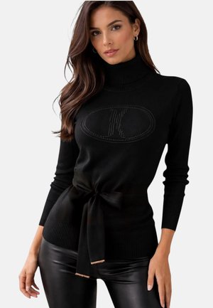 Donna con capelli castani lunghi che indossa un maglione nero a collo alto con fiocco frontale e pantaloni in pelle nera, in piedi su sfondo bianco.