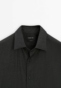 Chemise gris foncé à boutons avec fines rayures verticales et col classique, montrant la moitié supérieure et l'étiquette de la marque à l'intérieur du col.