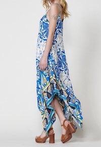 Femme portant une robe sans manches à motif bleu et blanc avec un ourlet asymétrique, des talons compensés beige et un tatouage en ligne sur le haut du bras.