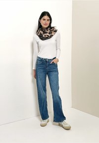 Haut blanc à manches longues, jeans évasés bleus, et une écharpe marron à motifs, associée à des baskets claires avec des accents beige et bleu.