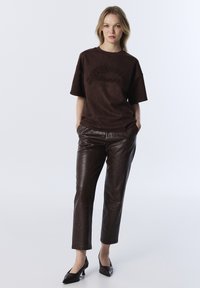 T-shirt marron surdimensionné avec texte en relief, associé à un pantalon en cuir marron ajusté avec un motif texturé. Des chaussures à talons noires complètent la tenue.