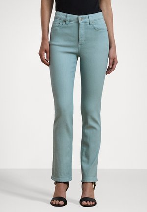 Femme portant un jean slim bleu clair et des talons ouverts noirs, debout devant un fond blanc uni.