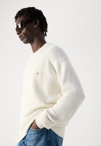 Man in tortoise shell zonnebril, witte gebreide trui met klein logo en blauwe jeans, poseert met één hand in zijn zak tegen een witte achtergrond.