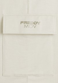 Tessuto beige caratterizzato da una tasca rettangolare con bordi cuciti. Sul fronte è riportato "FREDDY MOV" in un font metallico oro. Texture liscia.