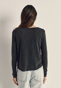 Pitkätukkainen nainen, jolla on tumma pitkähihaisen yläosa ja vaaleansiniset farkut, seisoo yksinkertaisen beigen taustan edessä selin.