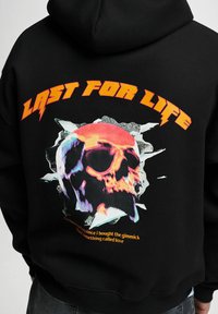 Fekete kapucnis pulóver, melyen színes koponya grafika található a "LAST FOR LIFE" felirattal, valamint további narancssárga szöveg alatta. Puha, texturált anyag.