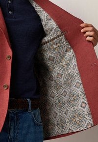 Roter Leinen-Blazer aus strukturiertem Stoff; blaue gefütterte Innenseite mit filigranen Mustern in Beige, Grün und Gold. Innentaschen sichtbar.