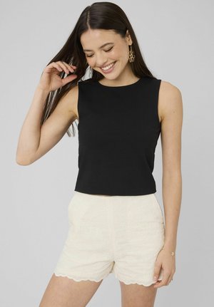 Femme souriante touche ses cheveux, portant un haut noir sans manches, un short crème texturé avec un ourlet festonné, et des boucles d'oreilles florales dorées devant un fond gris.