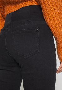 Jean en denim noir avec une taille haute, doté de poches arrière et d'une légère élasticité. La texture est lisse avec une couleur unie.