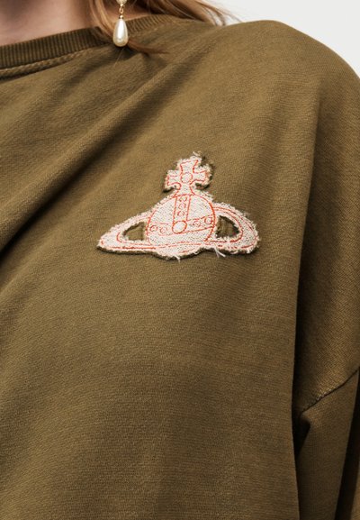 Ύφασμα ελιάς με σκληρό περιθώριο κεντημένου λευκού και πορτοκαλί πλανητικού συμβόλου με σταυρό στην κορυφή, κοντά στον ώμο.