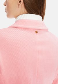 Rosa Cordjacke mit weißem geripptem Kragen, ausgestattet mit einem goldenen Akzentknopf auf der Rückseite und vertikaler Ripp-Textur.