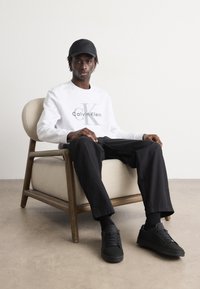 Felpa bianca Calvin Klein con logo grigio, pantaloni neri e sneakers nere, seduto su una sedia imbottita chiara. Sfondo neutro.