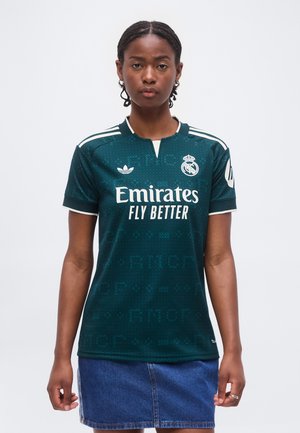 Femme portant un maillot de football vert foncé du Real Madrid avec le texte « Emirates Fly Better » et une jupe en jean bleue, debout au centre sur fond blanc.