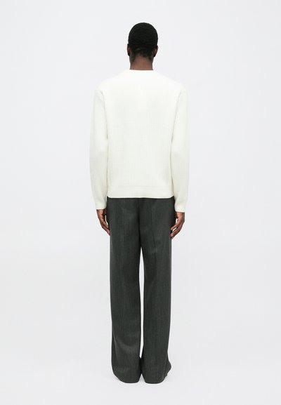 Man standing with back visible, wearing a white ribbed sweater and dark wide-leg trousers against a plain white background.