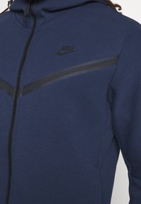 Navy blue zip-up hoodie made of soft fabric, featuring a black Nike logo and contrasting black stripes across the chest area.
