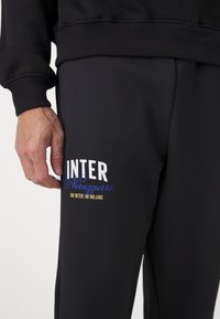 Crne trenirke s bijelim i plavim "INTER" logotipom te "Nerazzurri" natpisom u plavoj i zlatnoj boji, glatke teksture, uskog kroja.