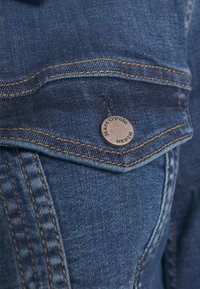 Detalj av en denimskjorta med en bröstficka och en cirkulär metallikon. Tyget är mörkblått med kontrasterande gula sömmar.