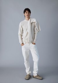 Maglione cremoso a maglia con motivo testurizzato, pantaloni bianchi e scarpe slip-on grigie; maglione appoggiato sulle spalle. Palette di colori neutri.