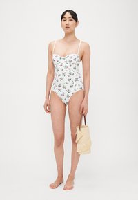 Costume intero bianco a fiori con spalline sottili, caratterizzato da fiori vivaci sul tessuto. La modella tiene in mano una piccola borsa intrecciata.