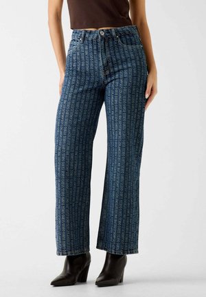 Donna che indossa jeans blu a gamba dritta con stampa verticale di testo bianco e stivali alla caviglia neri con tacco, in piedi contro uno sfondo semplice.