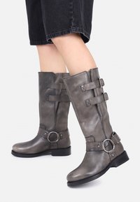 Botas de cuero gris hasta la rodilla con hebillas y anillas, llevadas con pantalones negros cortos sobre un fondo blanco.