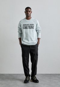 Světle modrý svetr s nápisem "Versace Jeans Couture" v černém písmu, spárovaný s černými cargo kalhotami s proužky a černými sportovními botami.