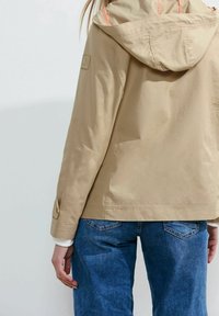 Beige vattentät jacka med huva, med orange detaljer och en logotyp-patch, i kombination med blå denimjeans. Slät, hållbar tyg.