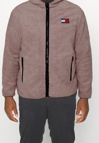Chaqueta de forro polar en rosa claro, con cremallera negra, dos bolsillos laterales y un parche de logo en rojo y azul en el pecho. Mangas largas con puños redondeados.
