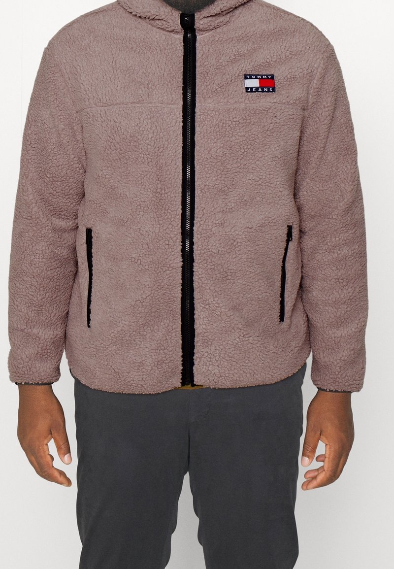 Chaqueta de forro polar en rosa claro, con cremallera negra, dos bolsillos laterales y un parche de logo en rojo y azul en el pecho. Mangas largas con puños redondeados.