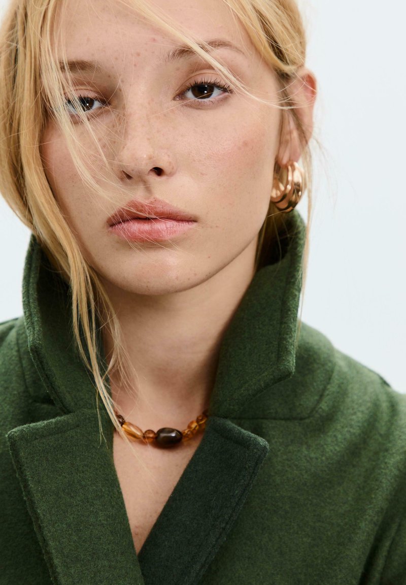 Zelený texturovaný kabát s vysokým límcem, v kombinaci s vícebarevným náhrdelníkem z korálků. Modelka má světlé vlasy a přirozený make-up.