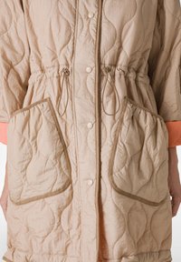 Abrigo acolchado de color beige claro con cintura ajustada, grandes bolsillos y detalles en las mangas en naranja contrastante. Presenta botones a presión y tejido textured.