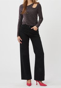 Schwarze weit geschnittene Hose kombiniert mit einem glitzernden dunklen Oberteil mit einem asymmetrischen Ausschnitt an der Schulter. Das Model trägt pinke High Heels.