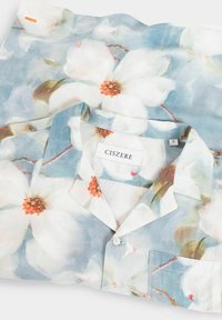 Ljusblå skjorta med ett blommönster av vita blommor med orange centrum, tillverkad av mjukt tyg, med krage och framlomm pockets.