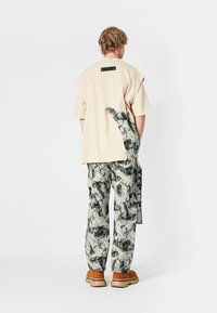 Persona con cabello rubio de espaldas, vistiendo una camisa beige de gran tamaño con un parche abstracto verde y unos pantalones a juego con un patrón en verde y blanco.