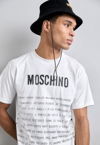 Hvit bomulls t-skjorte med fet svart "MOSCHINO" skrift og tekst på forskjellige språk. Korte ermer med rund hals.