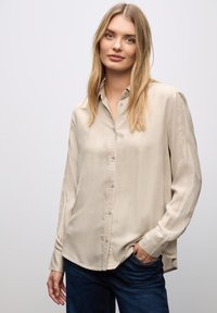 Camicia beige a maniche lunghe con bottoni realizzata in tessuto morbido, dotata di colletto classico e una sottile trama, abbinata a jeans in denim scuro.