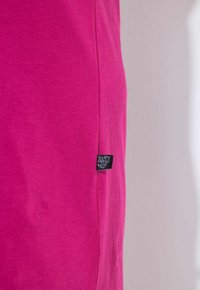 Felroze stof met een klein zwart ingenaaid label met witte tekst en decoratieve elementen nabij de naad.