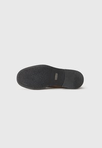 Suela de un zapato marrón que muestra un tacón de goma negra texturizada y el dibujo de la suela en la parte delantera, con una pequeña etiqueta rectangular de la marca en el medio.