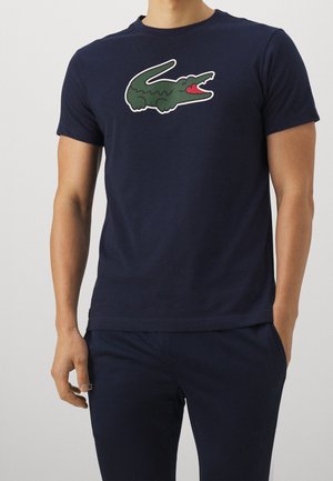 Mann trägt ein marineblaues T-Shirt mit einer großen grünen Cartoon-Krokodil-Grafik auf der Brust, kombiniert mit passenden marineblauen Hosen.