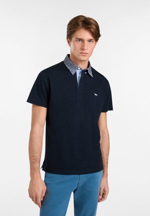 Mornarska polo majica s uzorkom na ovratniku, kratkim rukavima i malim logotipom na prsima. Tkanina izgleda glatko i teksturirano.
