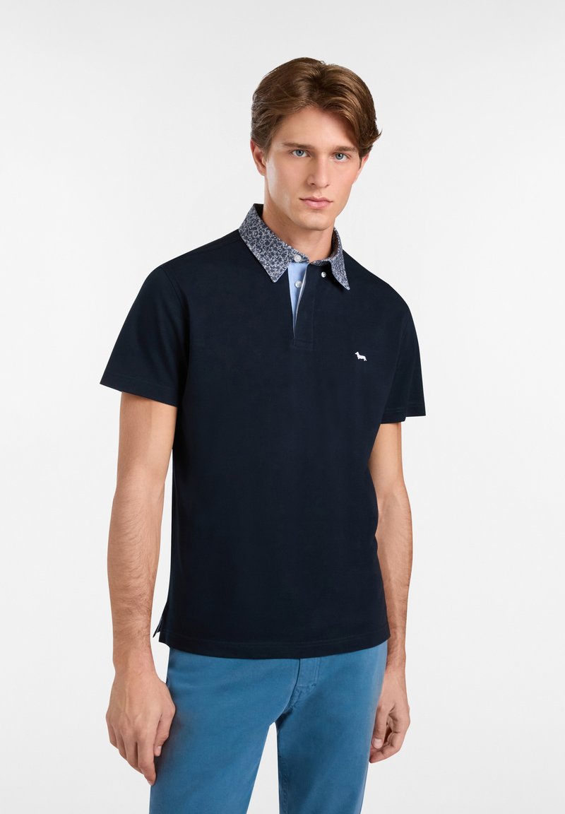 Námornícky polo tričko s vzorovaným golierom, krátkymi rukávmi a malým logom na hrudi. Materiál sa zdá byť hladký a textúrovaný.