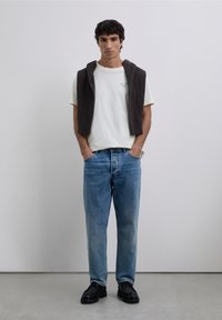 Weißes T-Shirt mit Logo, brauner Pullover über den Schultern drapiert, blaue Jeans und schwarze Schuhe, stehend vor einem schlichten Hintergrund.