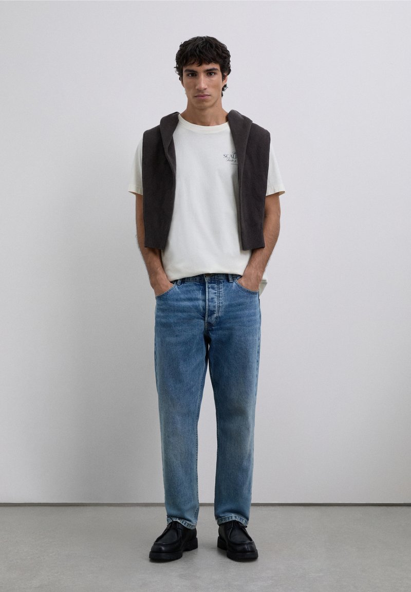 Weißes T-Shirt mit Logo, brauner Pullover über den Schultern drapiert, blaue Jeans und schwarze Schuhe, stehend vor einem schlichten Hintergrund.