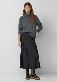 Pull gris à col roulé, associé à une jupe en satin noir et des bottes à plateforme noires. Le pull présente des poignets côtelés et des boutons sur le côté.