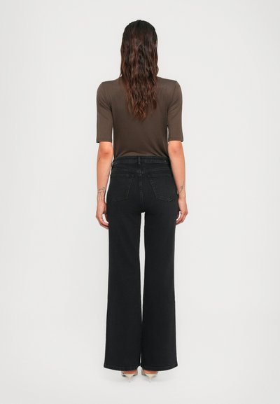 Woman with long wavy hair wearing a brown short-sleeve top, high-waisted wide-leg black jeans, and beige heels, standing facing away.